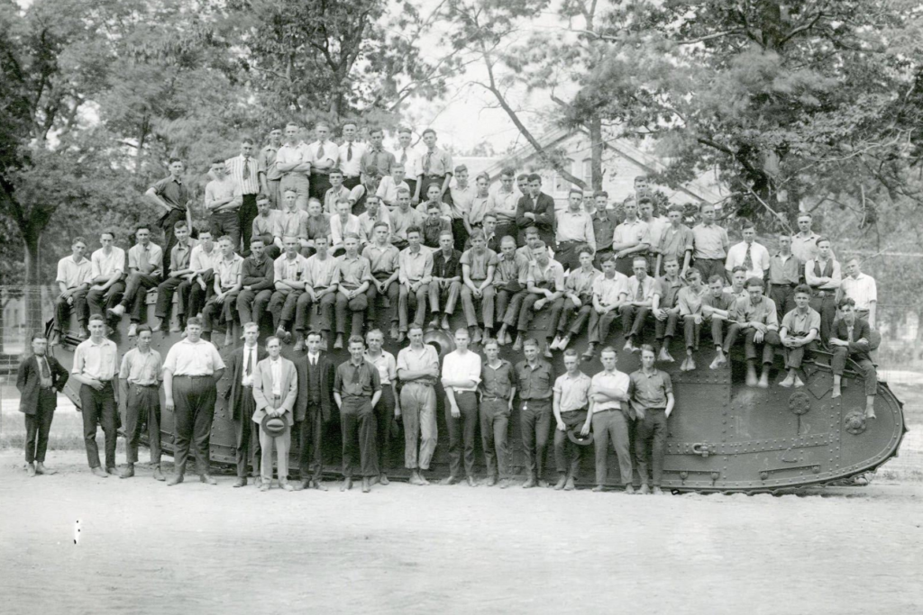 Black and white photo of a group of approximately 50 men sitting on and standing in front of a Mark VIII Liberty Tank in 1920.