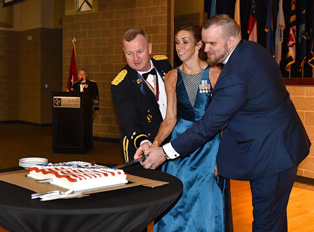 A military officer in dress uniform, a woman in a teal gown, and a young man in a navy suit cut a sheet cake decorated with a red-and-white striped design at a formal event, with an officer speaking at a "Waterfront Convention Center" podium and rows of flags in the background.