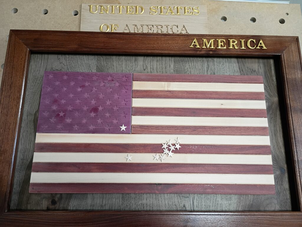 A handcrafted wooden American flag artwork in a dark wood frame, with the stripes carved from alternating light and dark wood and the star field left unfinished, small metal star pieces scattered loose on the stripes; a separate wooden sign reading "United States of America" leans behind it, and "AMERICA" is engraved in gold on the frame.